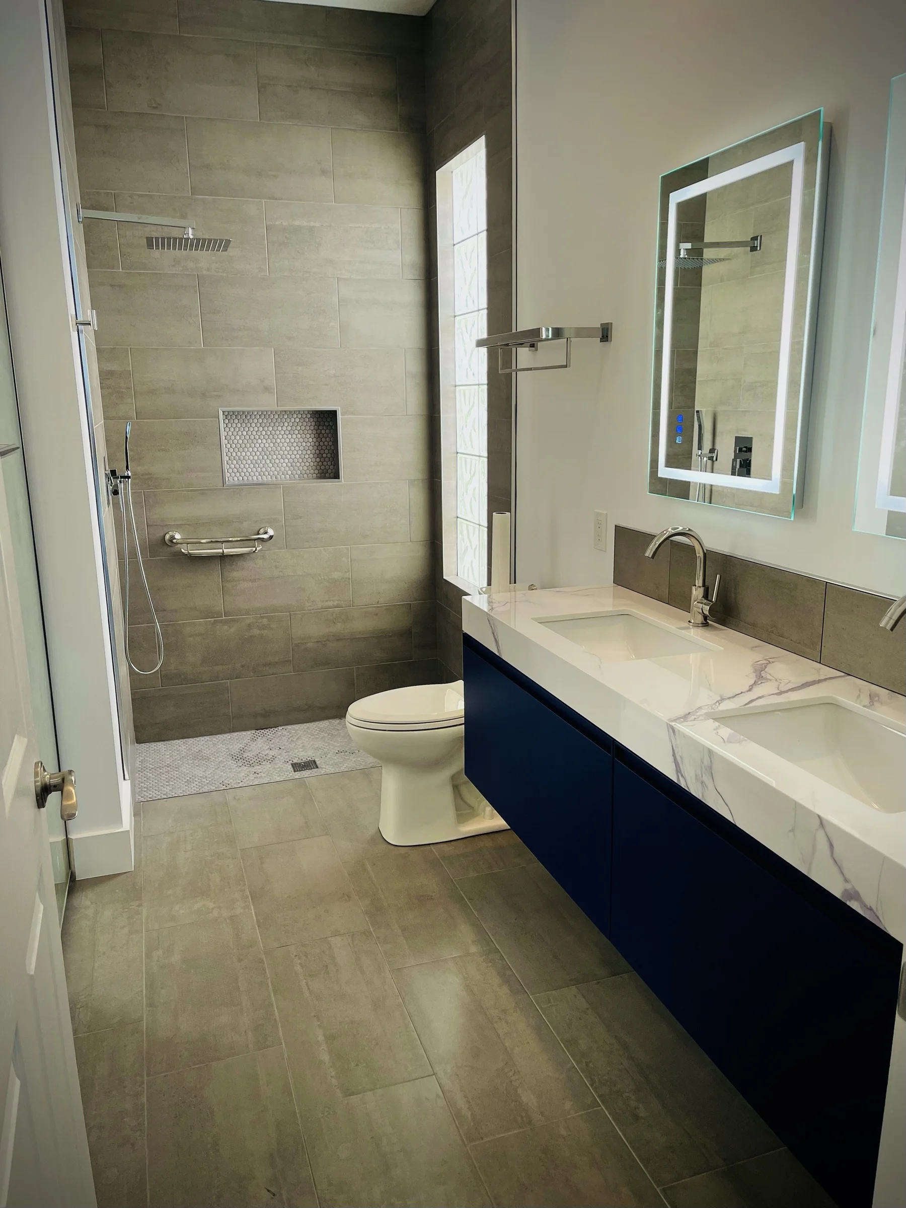Finished bathroom with a gray tiled walk-in shower, navy double vanity, stone counter, and illuminated mirrors
