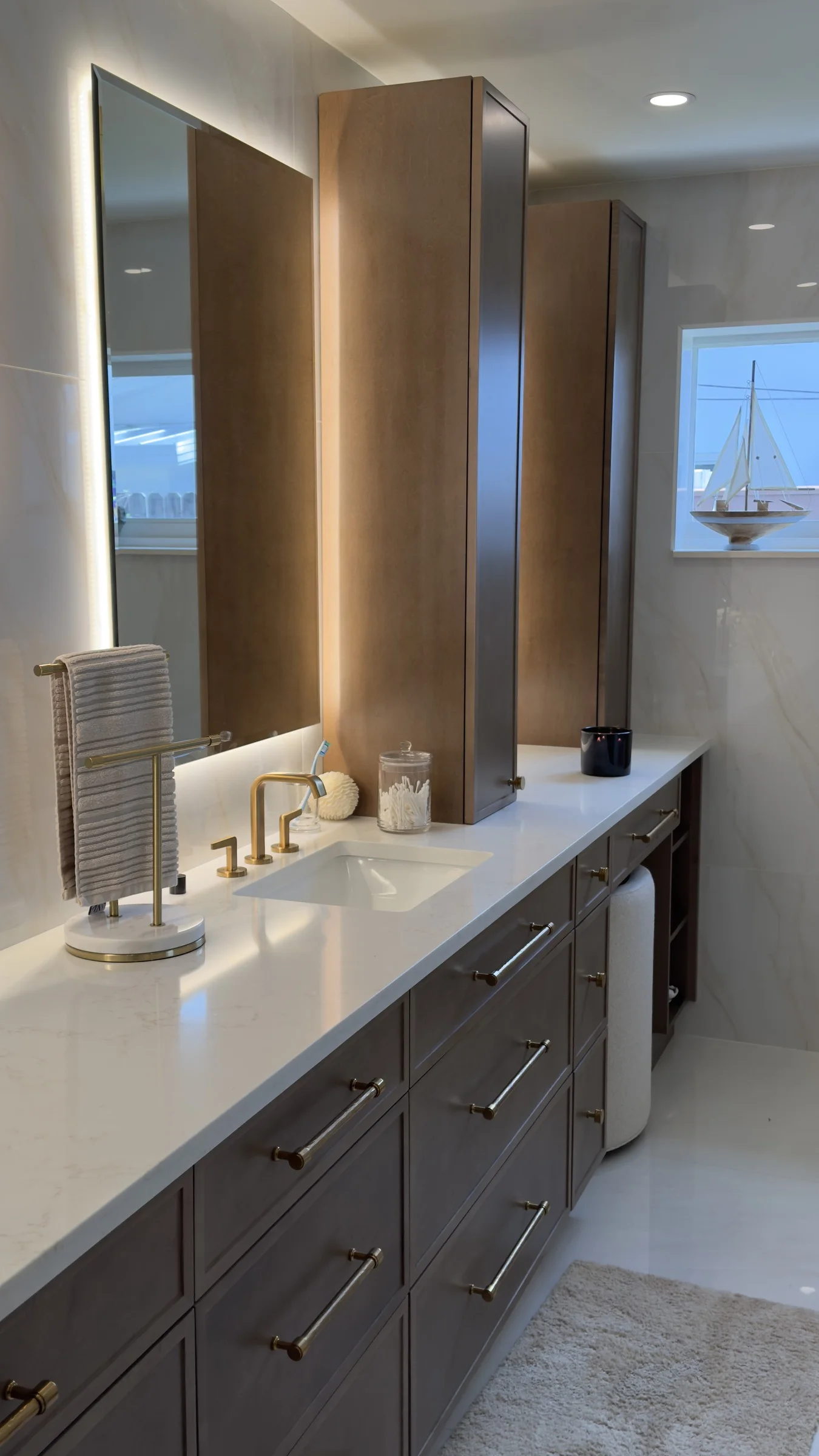 Long dark wood bathroom vanity with a white countertop, brass faucets, and illuminated mirrors
