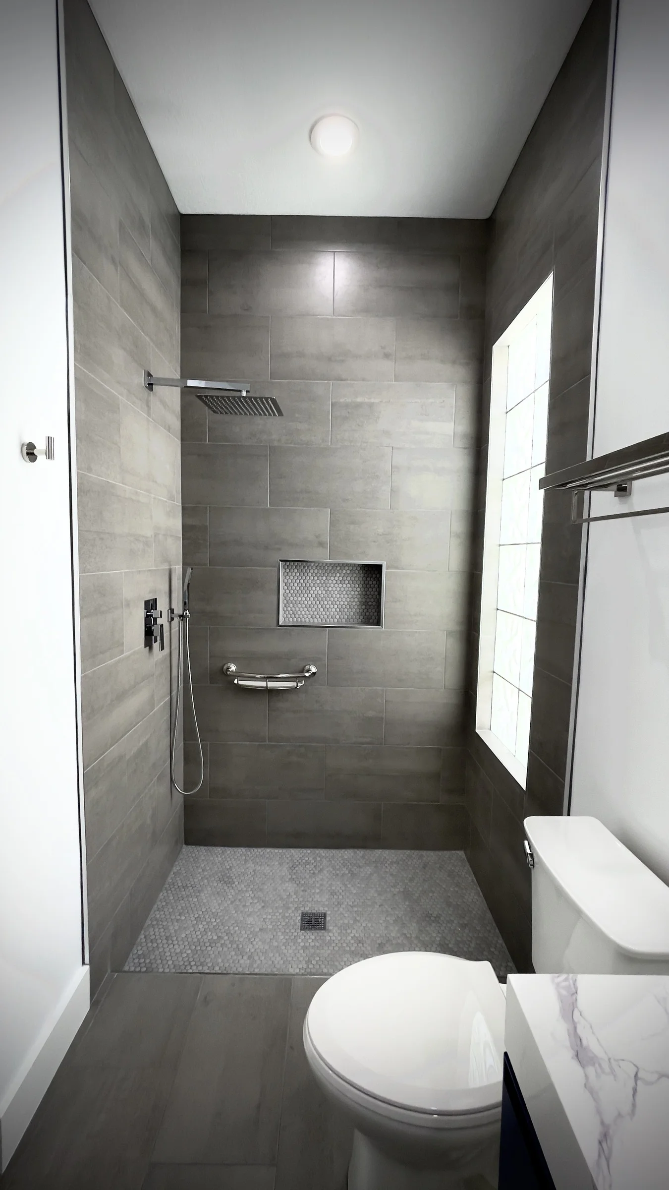 Gray tiled walk-in shower with a built-in niche, rain shower, handheld fixture, and grab bar