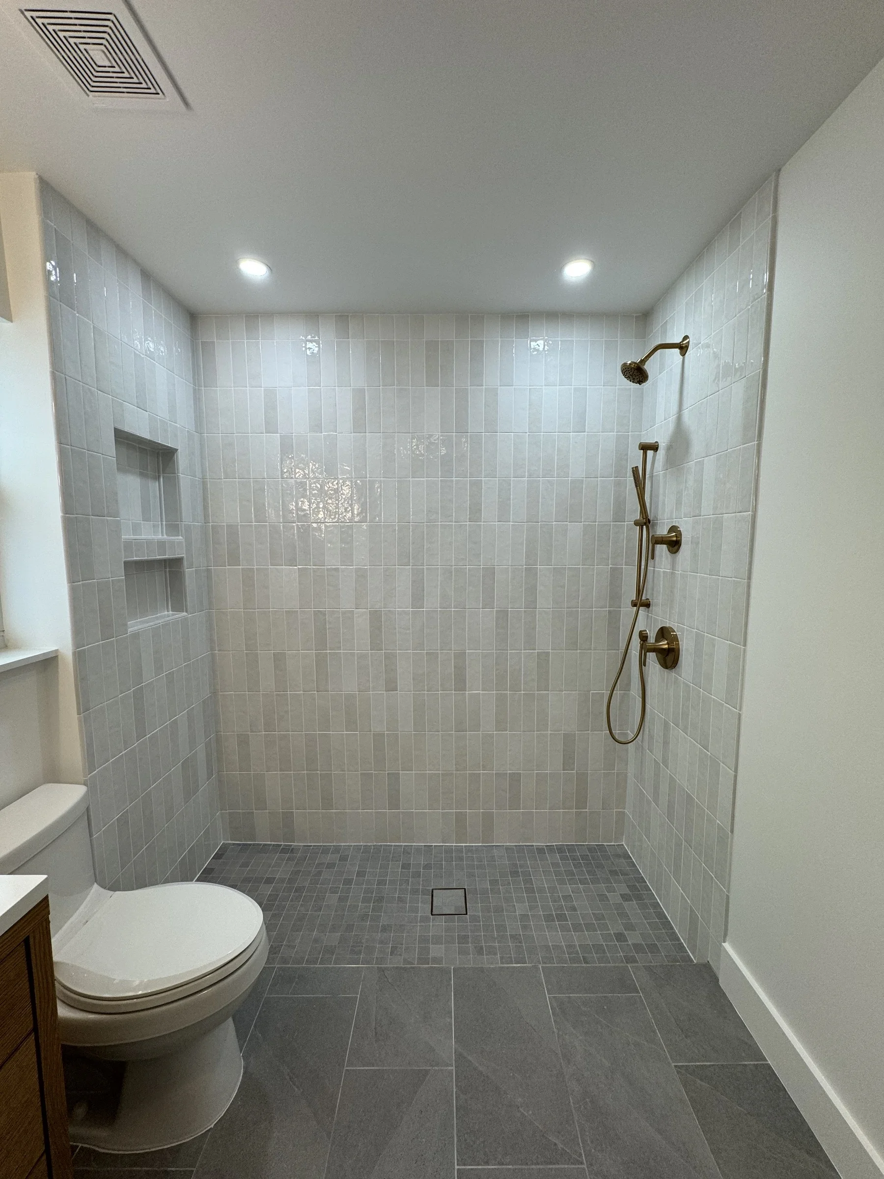 Finished walk-in shower with pale tile, gray mosaic floor, a built-in niche, and brass fixtures