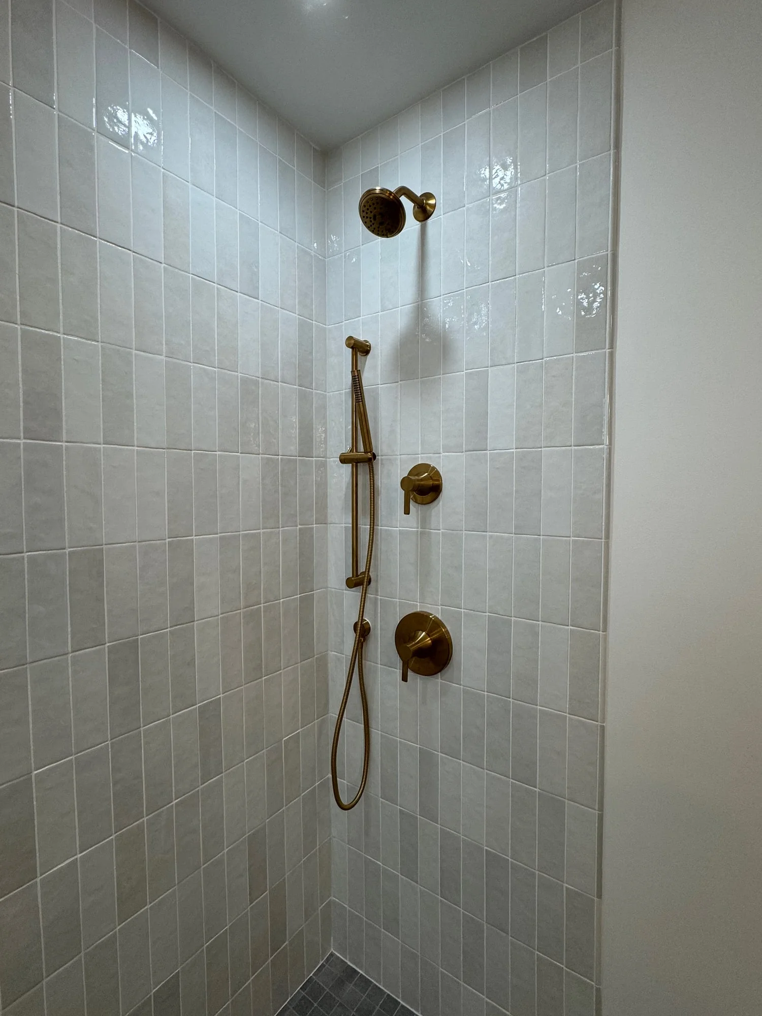 Brass-colored showerhead, handheld fixture, and controls against pale vertically laid shower tile