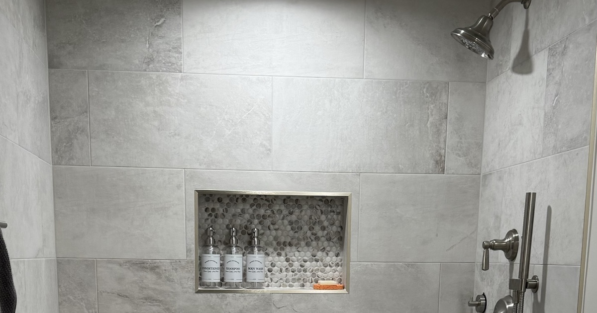Landscape detail of a tiled stand-up shower showing the shower niche, showerhead, and controls.